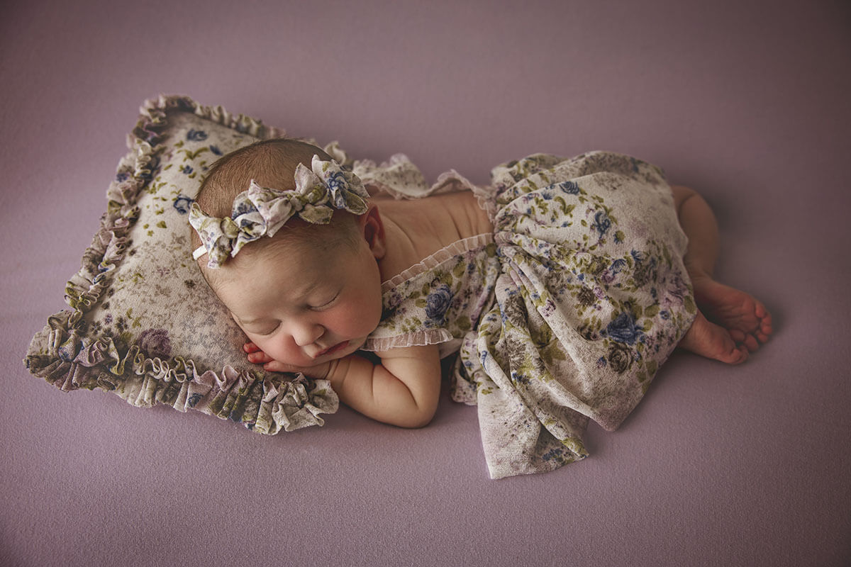 A newborn baby girl in a floral pattern dress with matching head band and pillow sleeps on her tummy on a purple bed after meeting lactation consultants in bloomington il