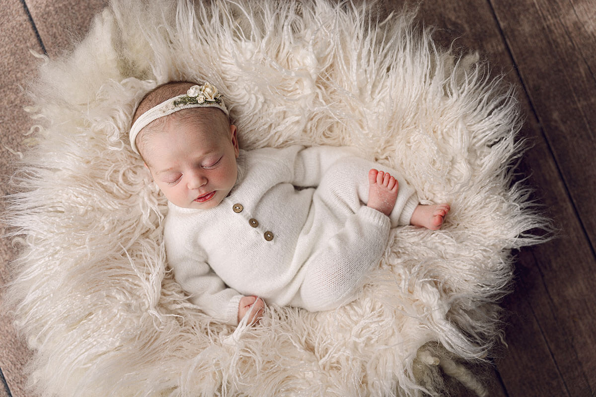 A newborn baby girl in a white onesie and matching flower headband sleeps on a shag pillow after meeting lactation consultants in bloomington il