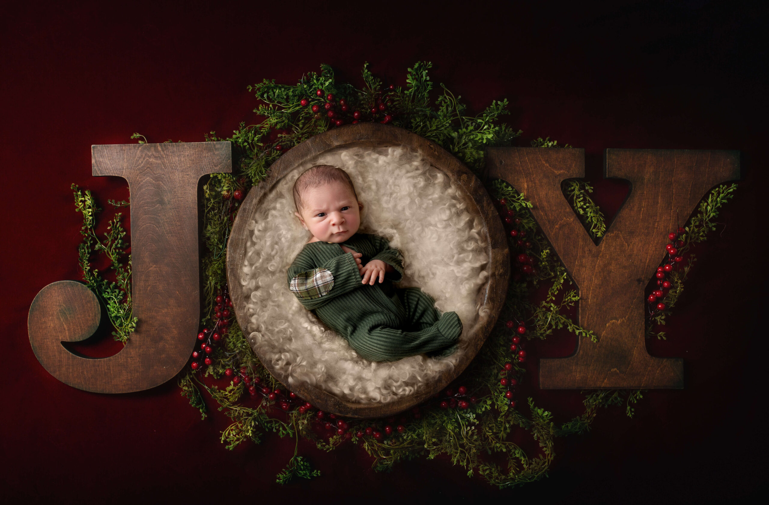 A grumpy newborn baby laying in the O of a wooden Joy in a green onesie thanks to an obgyn in bloomington il