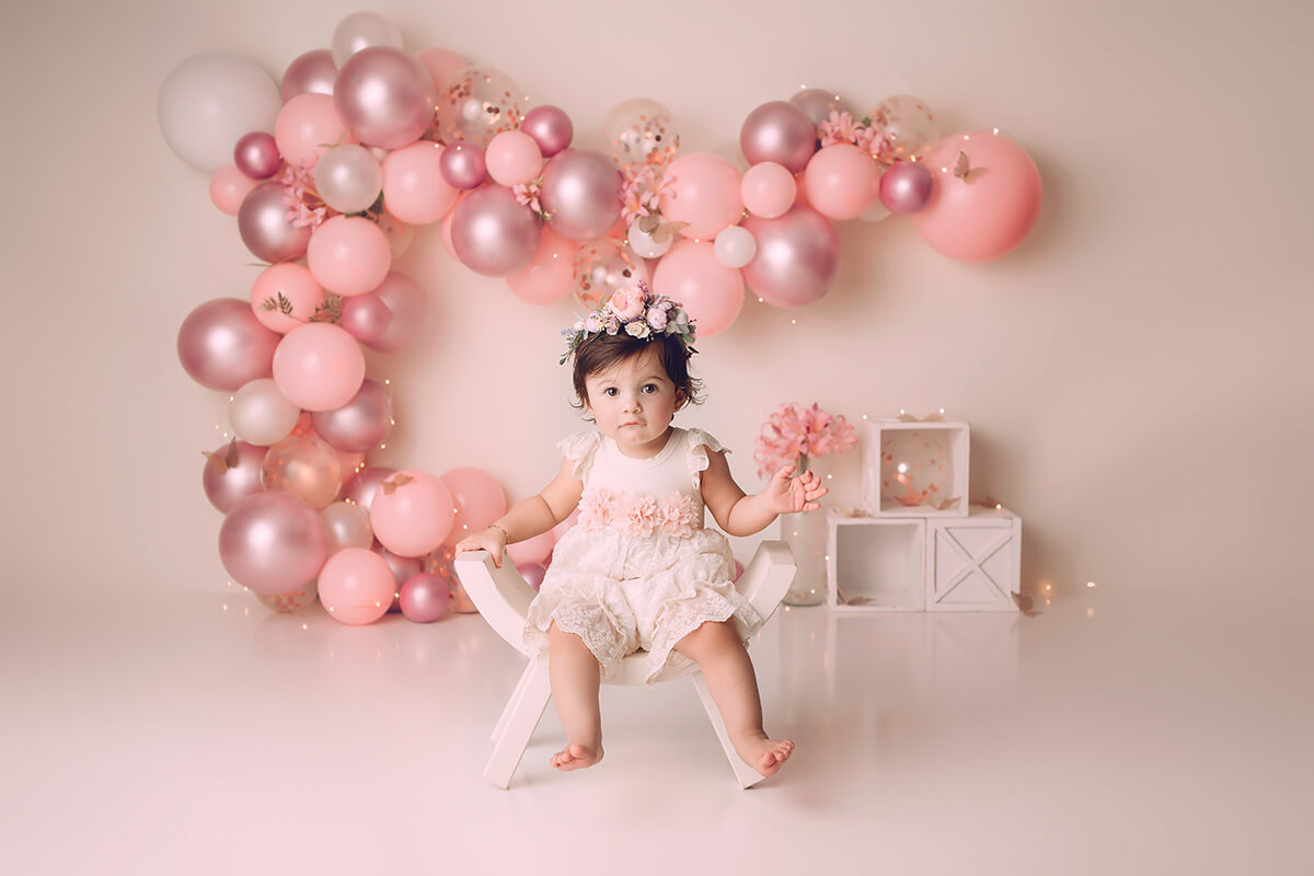 A young girl in a pink dress and matching flower head piece sits on a curved wooden stool in a studio with pink balloons on the wall