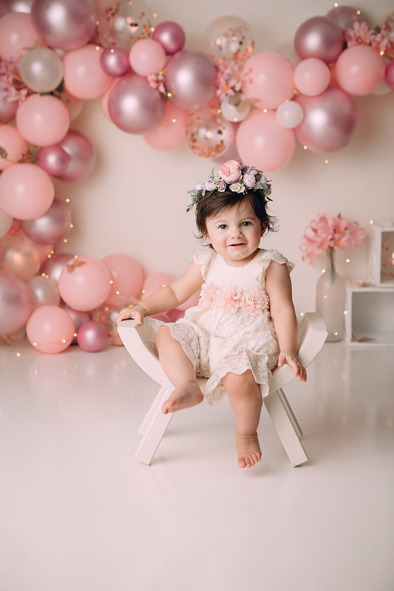A happy toddler girl in a pink dress sits on a wooden bench in a birthday themed studio