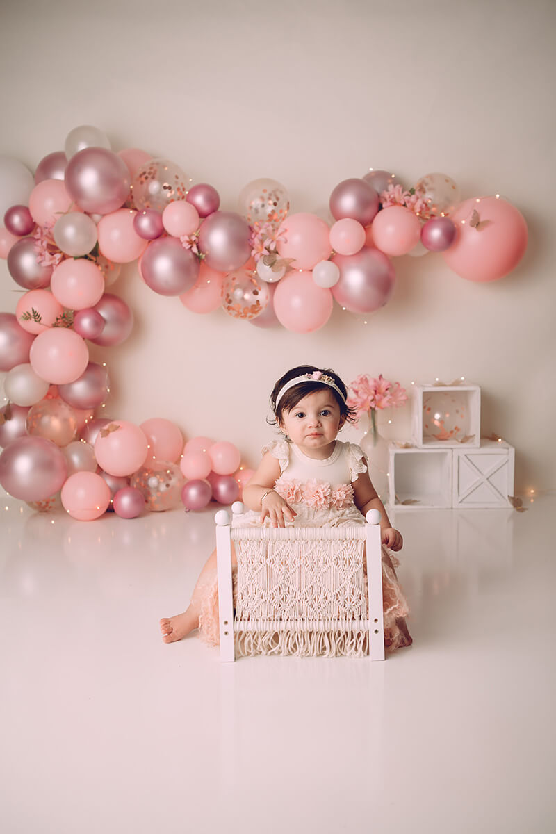 A toddler girl straddles a tiny bed in a studio with pink balloons on the wall after visiting pediatric dentists in bloomington il