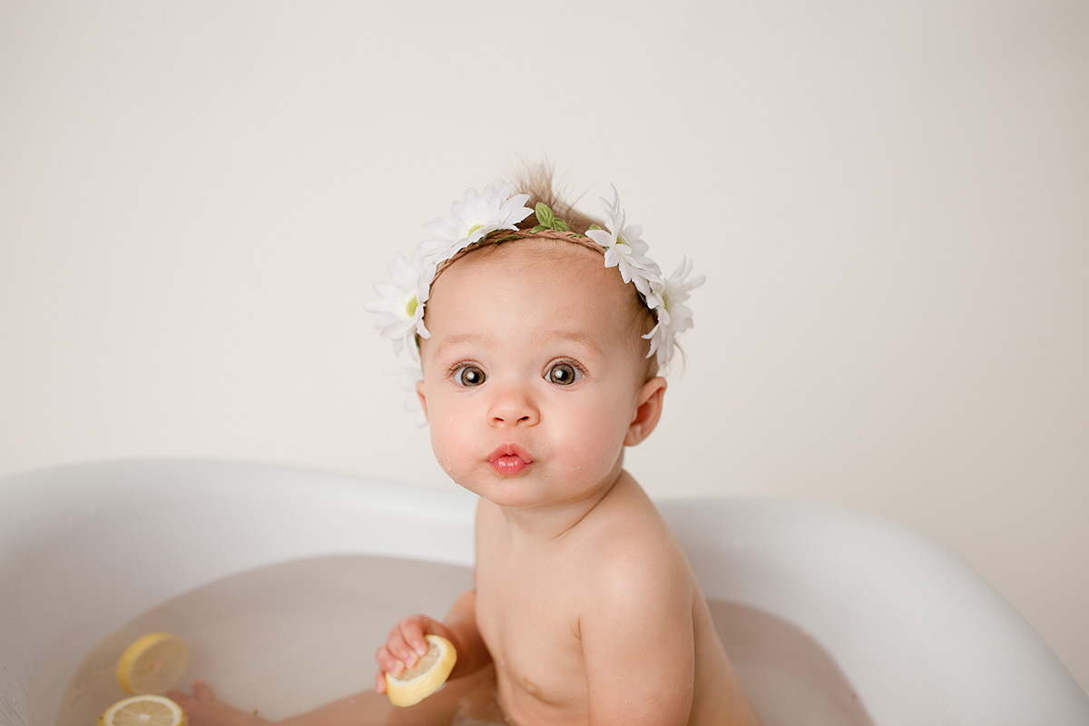 A baby girl in a white flower headband holds a lemon slive while sitting in a tiny bathtub after meeting pediatricians in bloomington il
