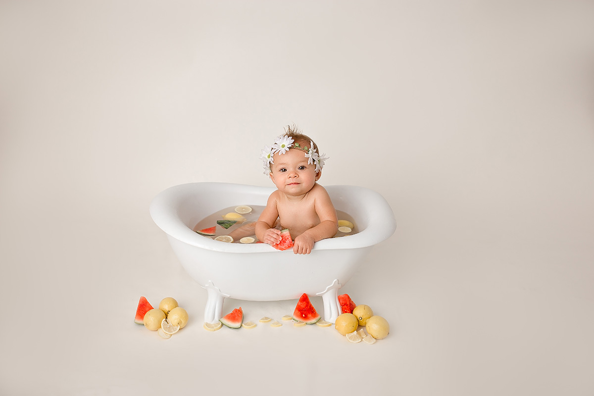 A smiling baby with a flower headband leans on the edge of a tiny bathtub filled with fruit after meeting pediatricians in bloomington il