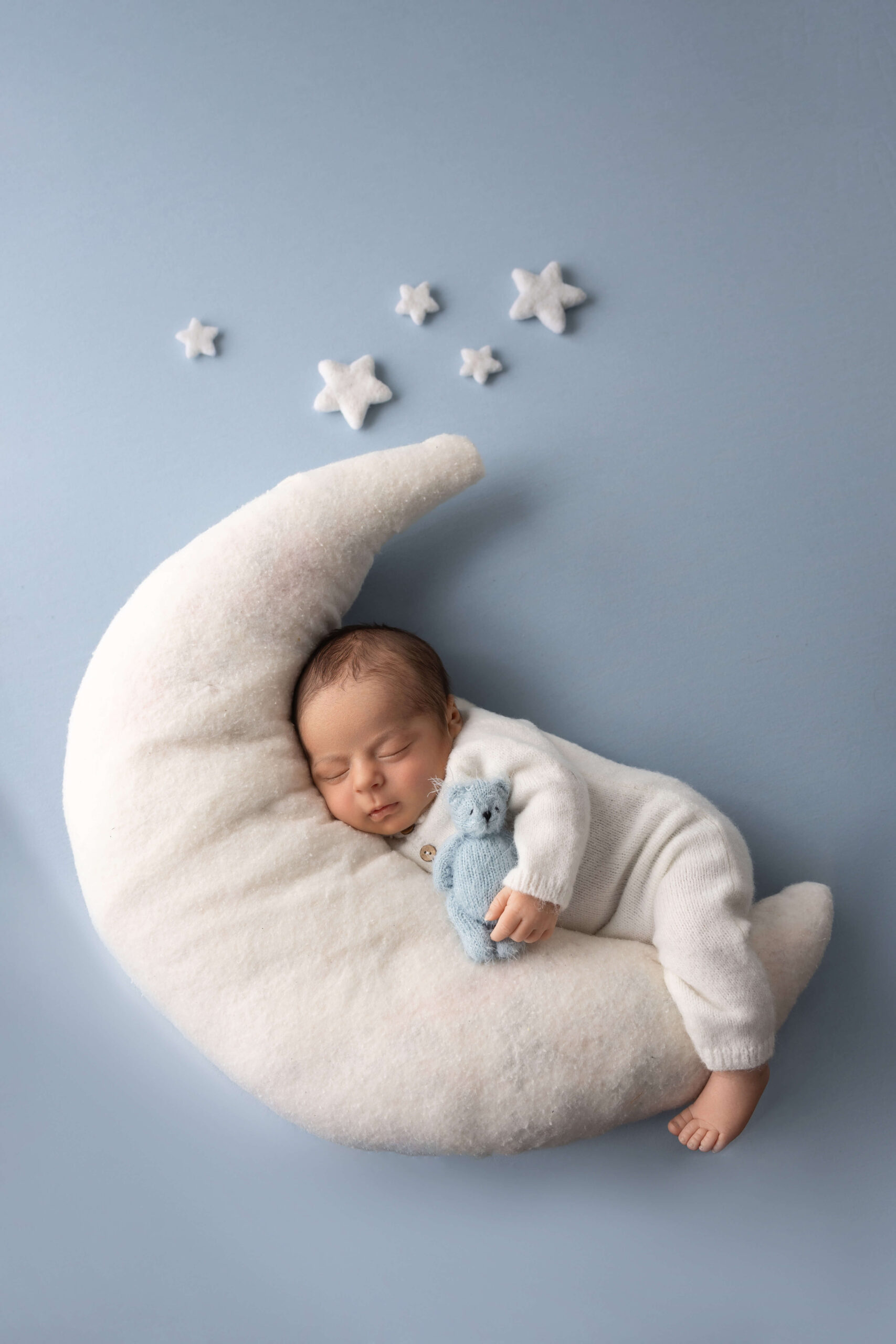 sleeping newborn in a white onesie hugging a blue teddy on a stuffed moon