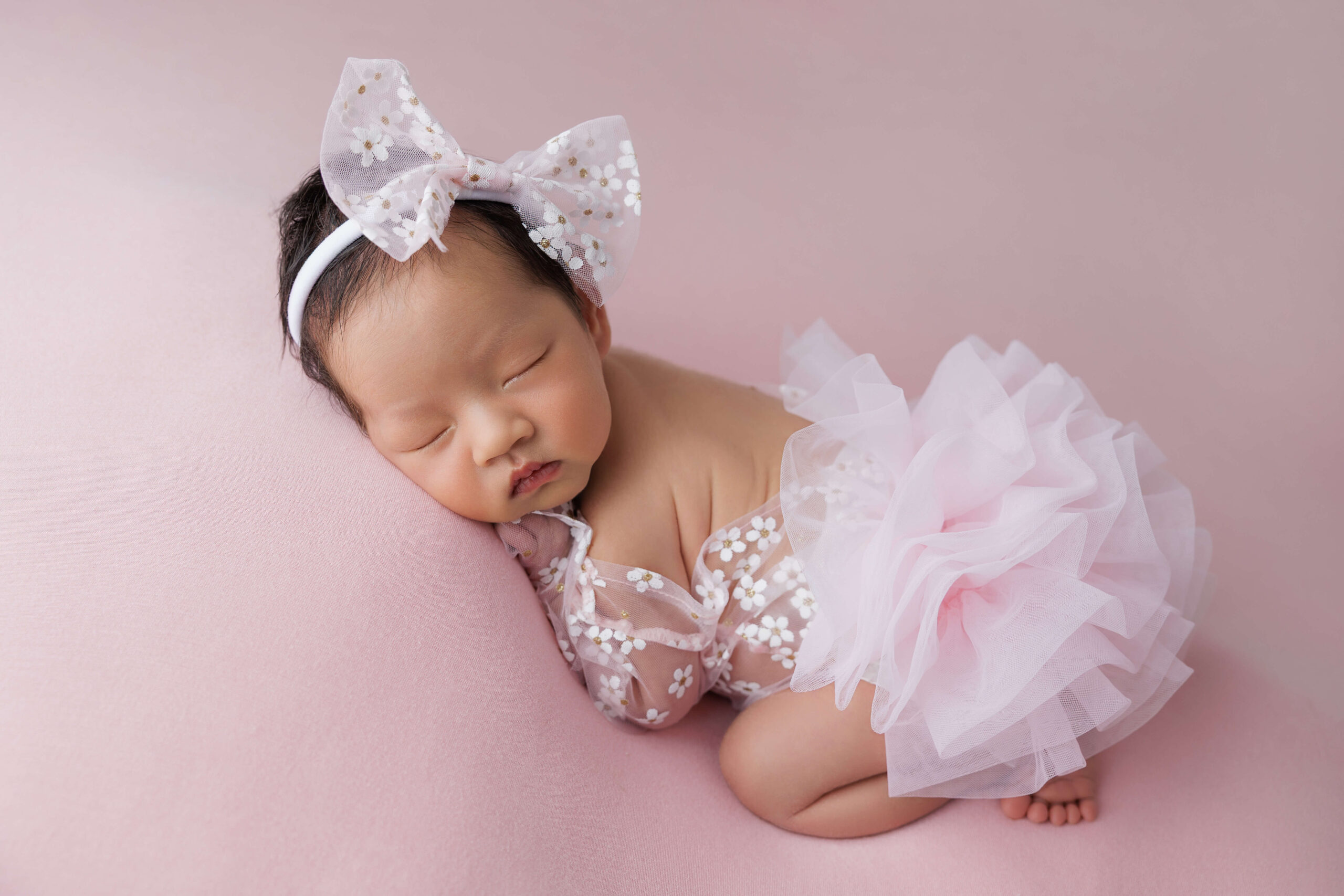 A newborn girl in a pink lace gown sleeps on her tummy with a large batching bow