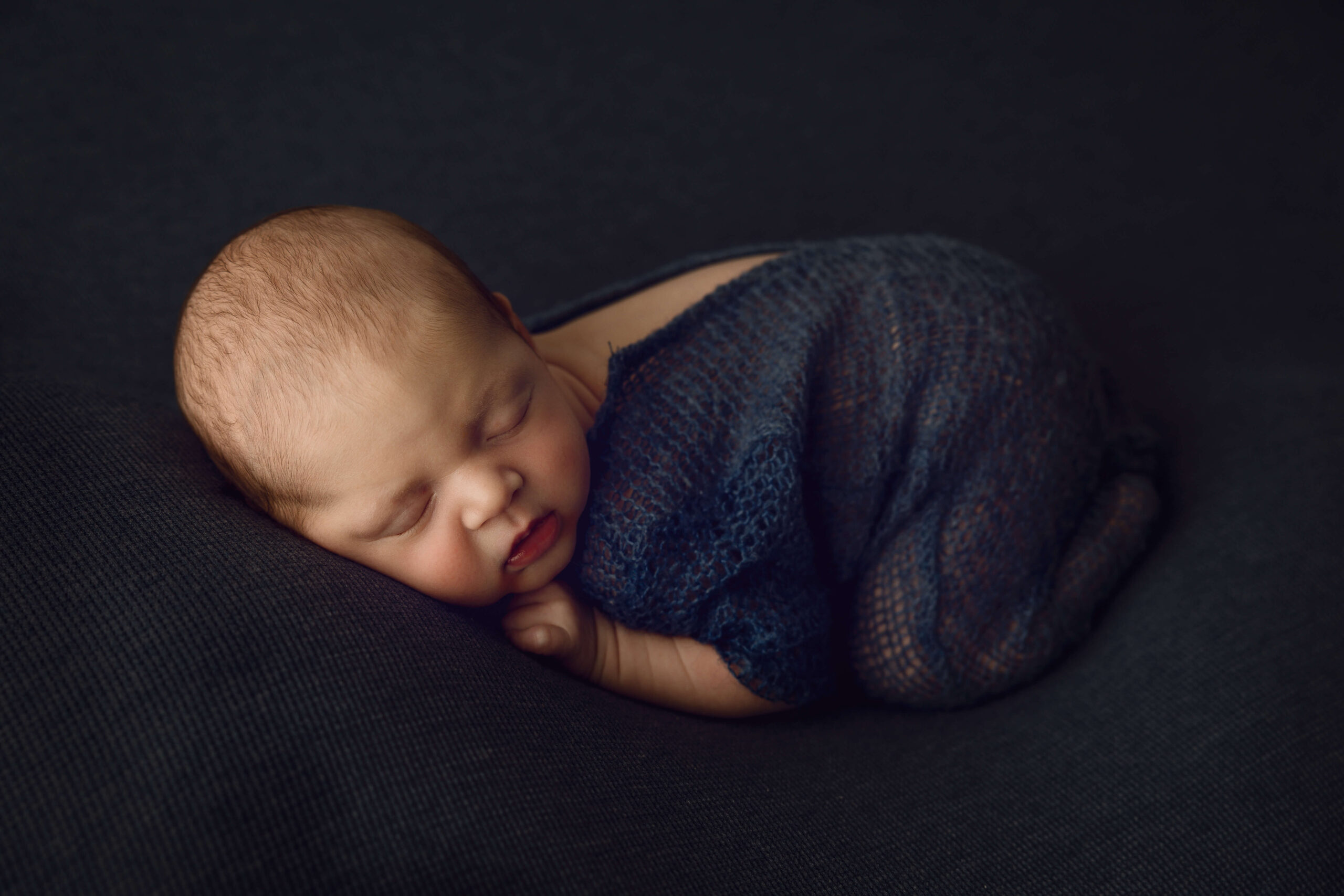 A newborn baby in a knit blue onesie sleeps in froggy pose in a studio