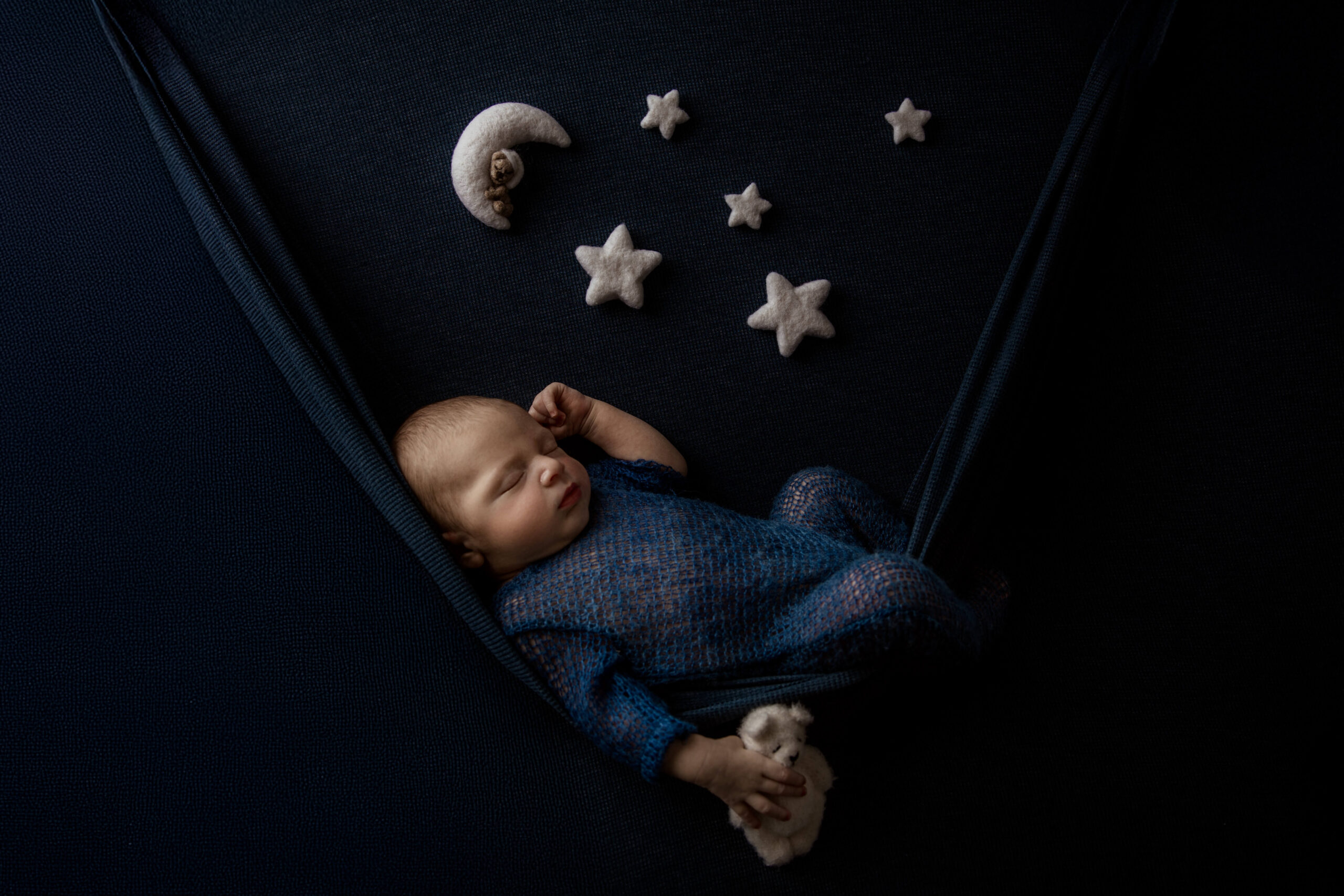 A sleeping newborn baby in a knit blue onesie under felt stars on a hammock after mom enjoyed a prenatal massage in bloomington il