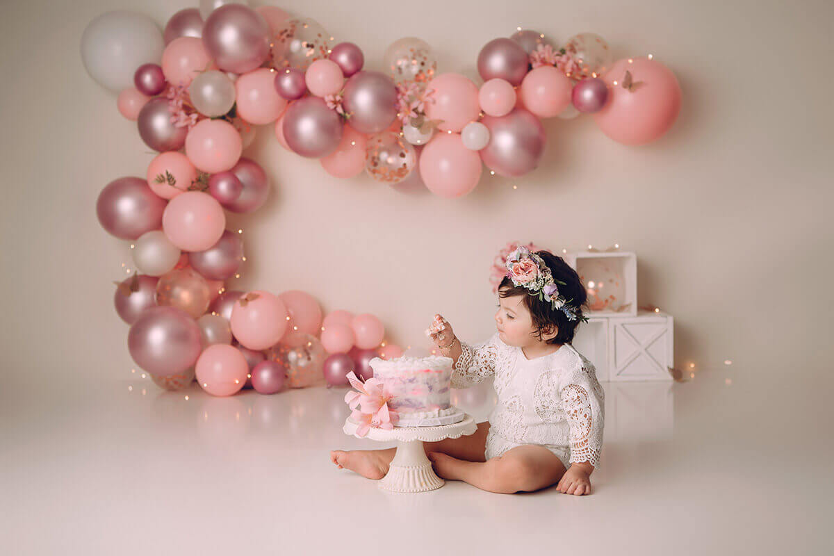 A happy baby girl sits in front of a pink balloon wall decor eating a cake with her hands in a white lace onesie