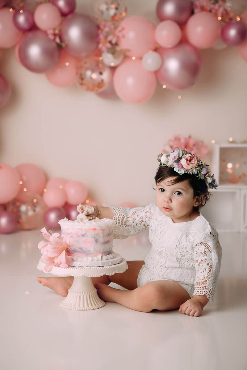 A baby girl in a flower headband and lace onesie sits with a hand in her birthday cake from one of the bakeries in springfield il