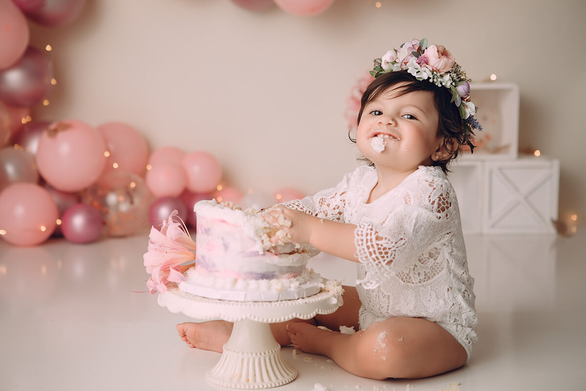 A happy baby girl smiles while eating a birthday cake in a lace onesie and flower headband
