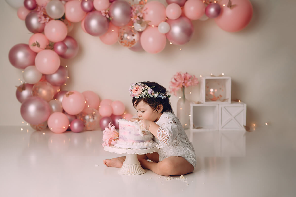A toddler girl digs into a pink cake in a studio in a white lace onesie during one of her birthday parties in Bloomington, IL