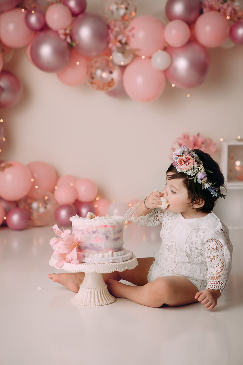 A young toddler takes a bite out a birthday cake while sitting in a studio in a lace white onesie