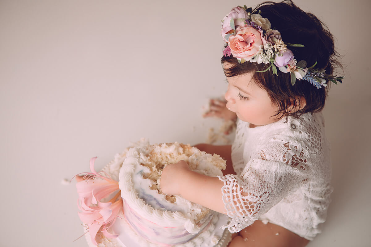 A look down to a baby girl smashing a cake in a white lace onesie and flower headband during one of her birthday parties in Bloomington, IL
