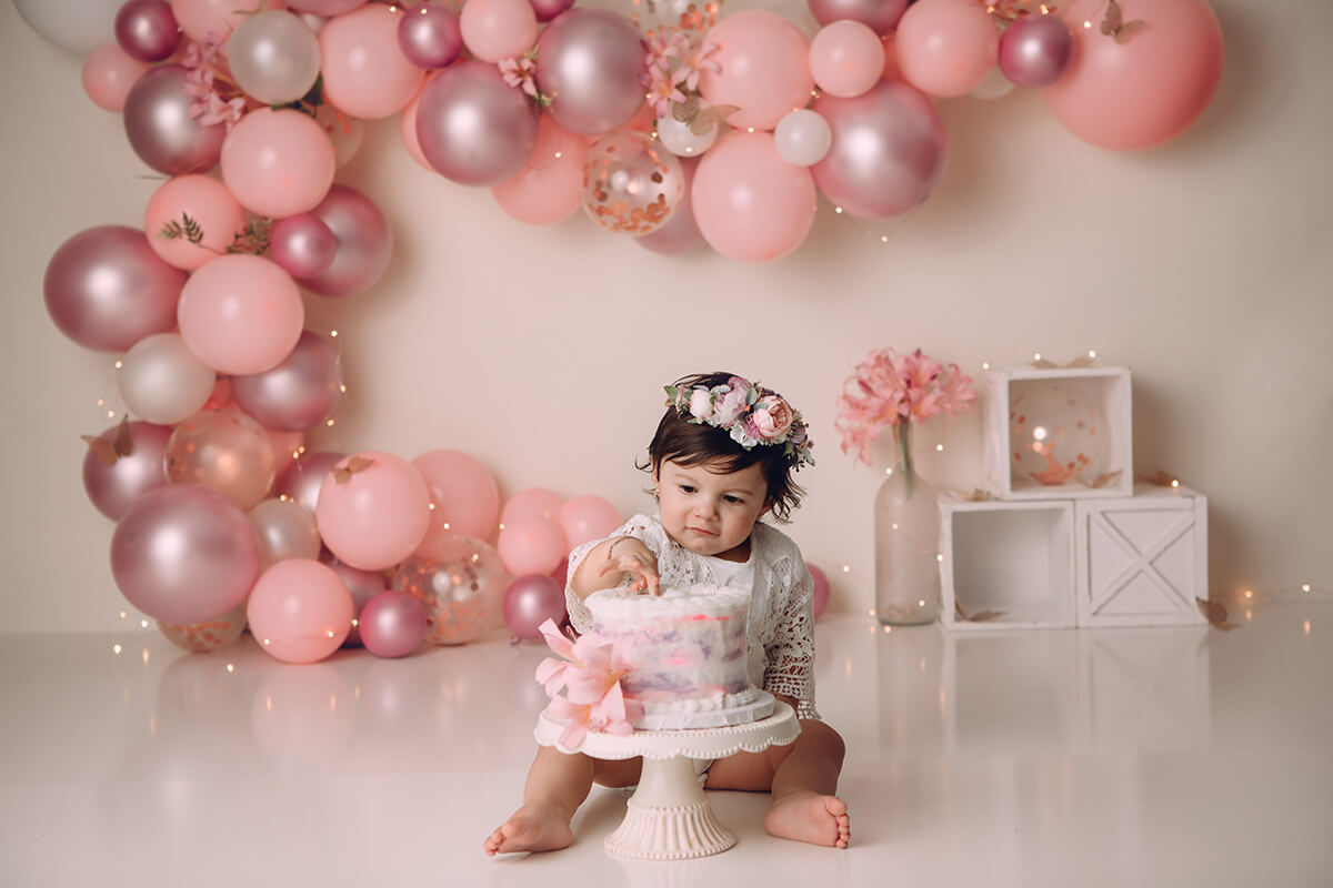 A toddler girl in a white dress sits with a flower headband reaching for a birthday cake in a pink studio during her birthday parties in Bloomington, IL