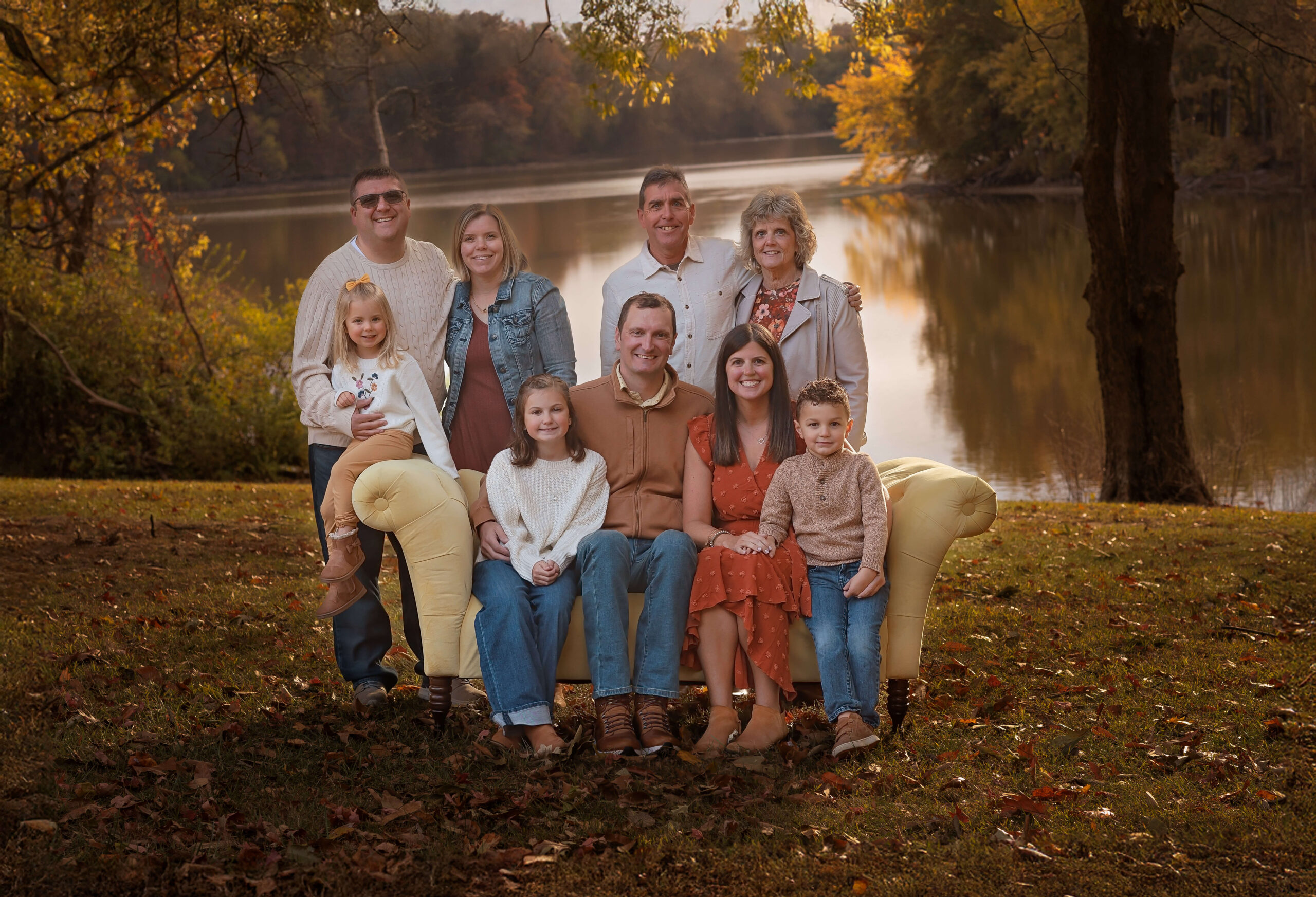 A happy family sits and stands around a couch in a park lawn at sunset by a lake after exploring the children's discovery museum