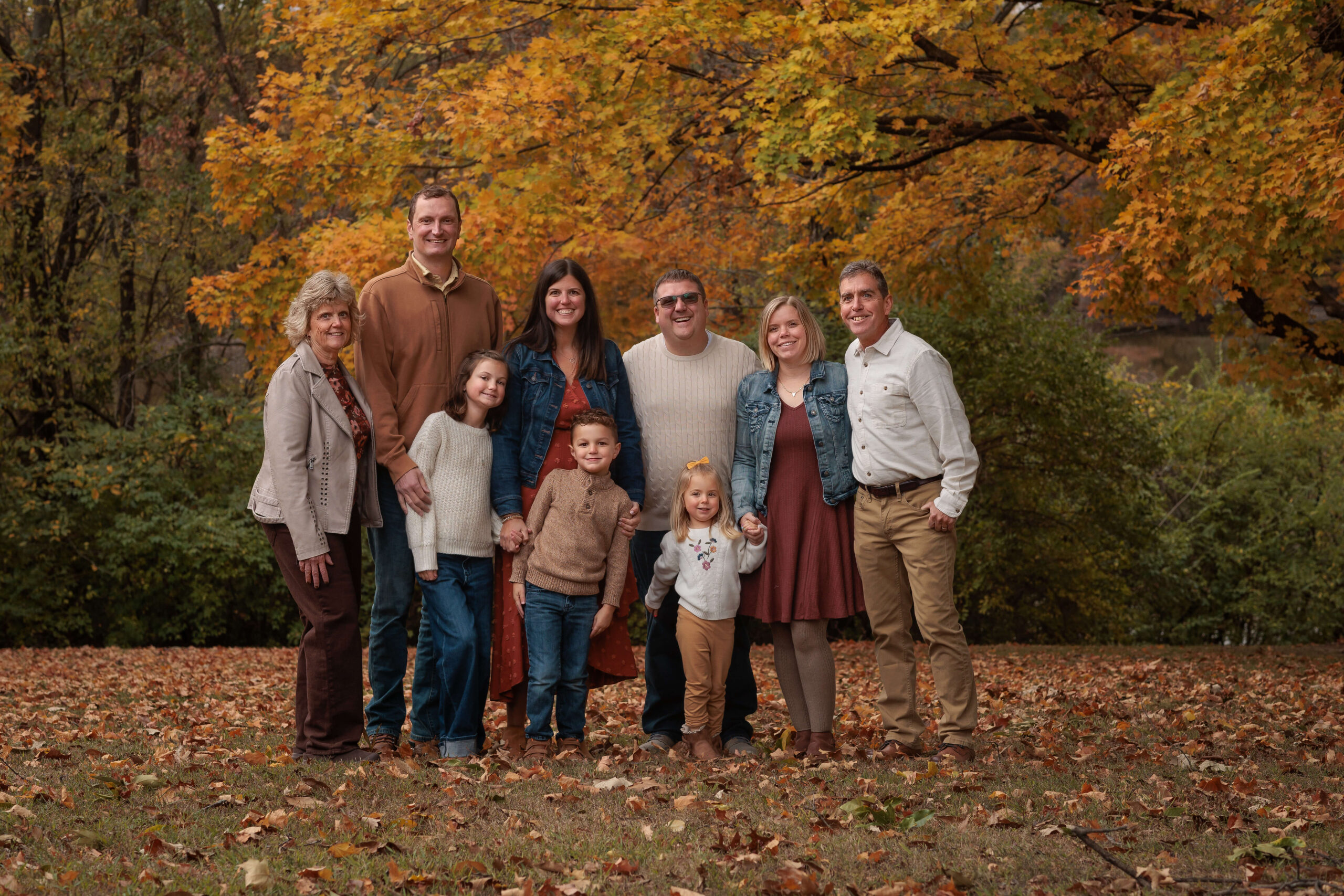 A smiling extended family with three little ones stand under fall color trees in a park lawn