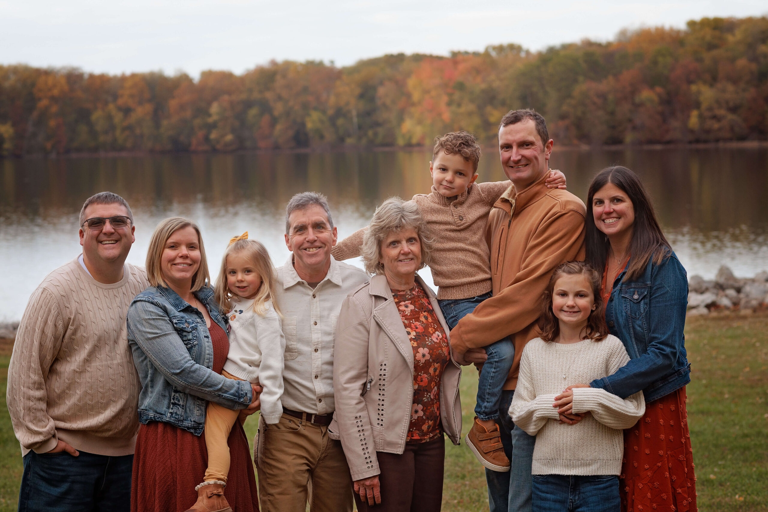 A happy extended family stands smiling surrounding grandma and grandpa in a park by a lake in fall after exploring children's discovery museum