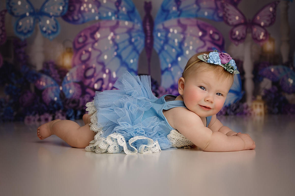 A baby girl lays across the studio floor in a blue gown and matching flower headband