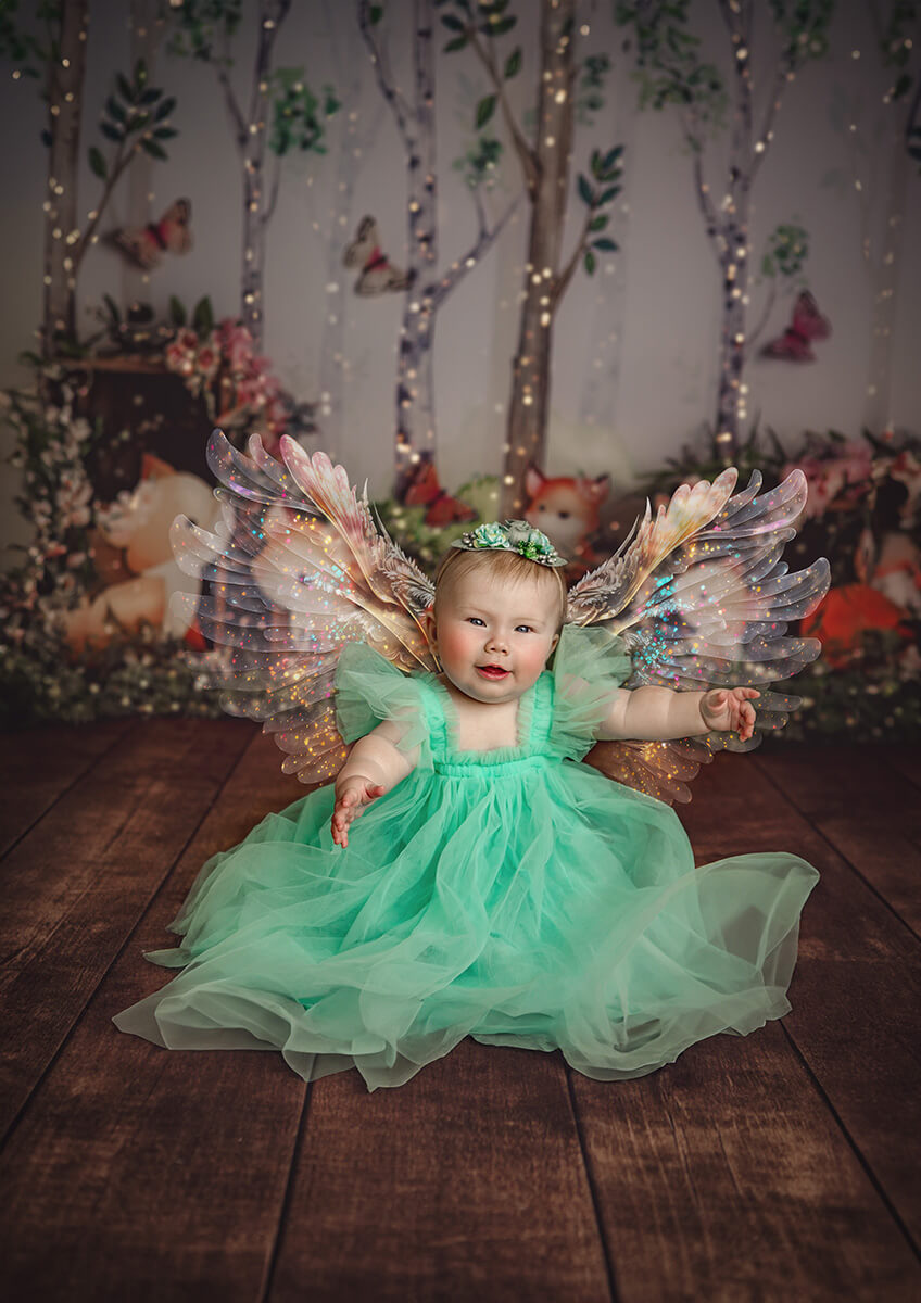 A baby girl sits in a green gown with large angel wings in a studio while wearing daisy diapers