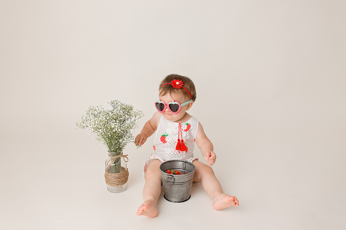 A baby girl plays with a bucket and wildflower in strawberry sunglasses and matching white onesie