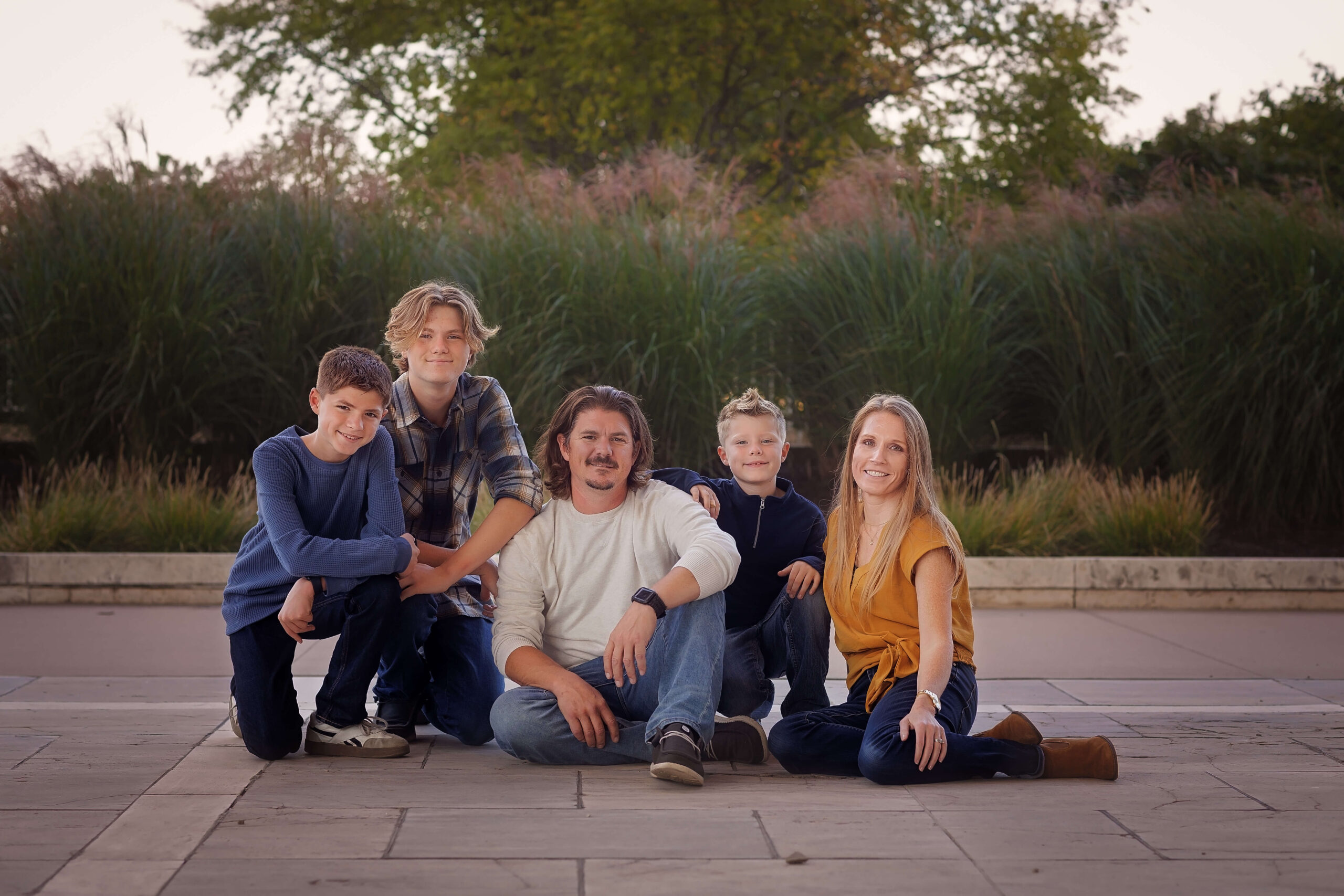 A family of five sit and kneel in a park sidewalk in front of tall pompous grass at sunset