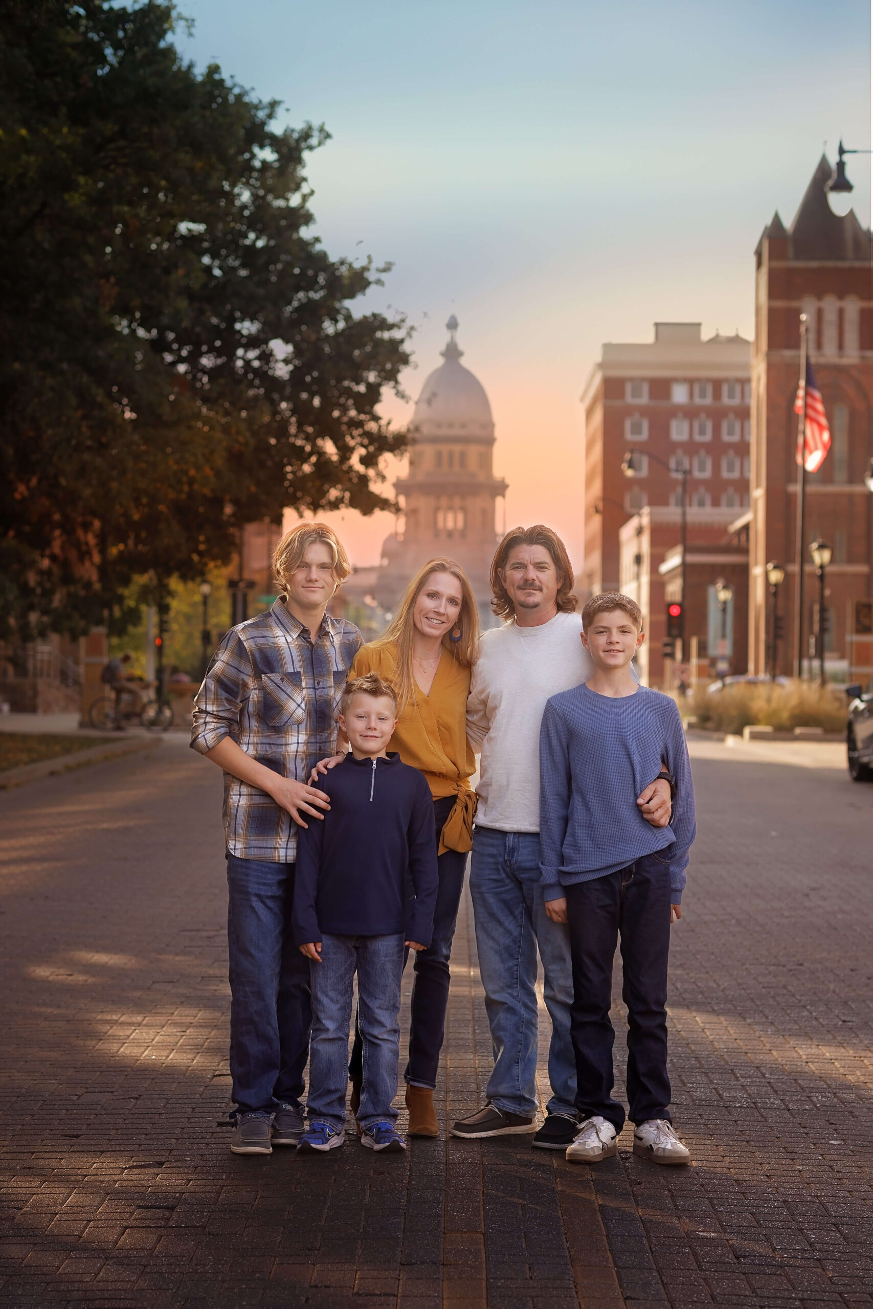 A mom and dad stand smiling with their three teen sons in a downtown brick sidewalk at sunset thanks to family counseling in bloomington il