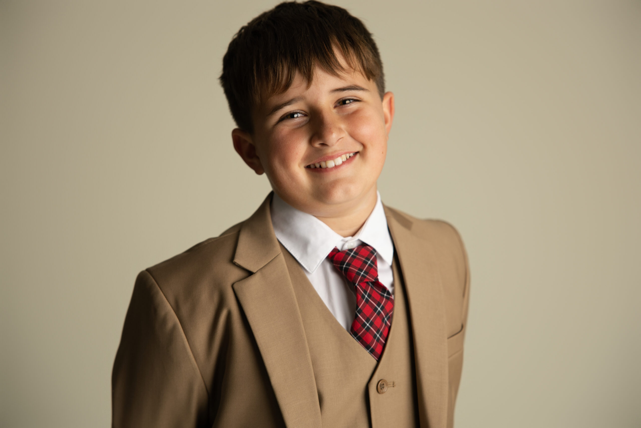 A young boy smiles while standing in a tan suit and red plaid tie