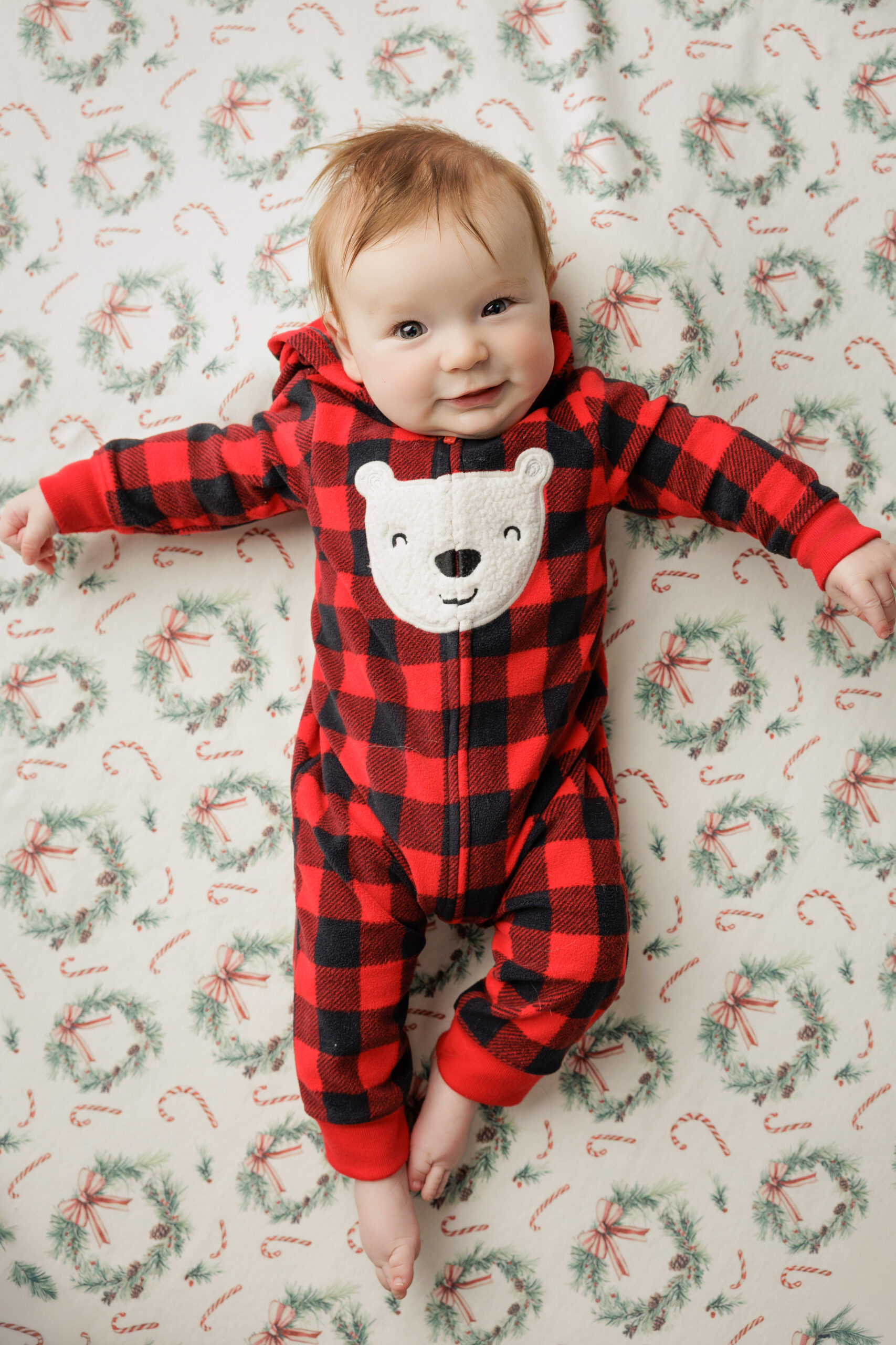 An infant laying in red plaid pajamas with eyes open and arms stretched out