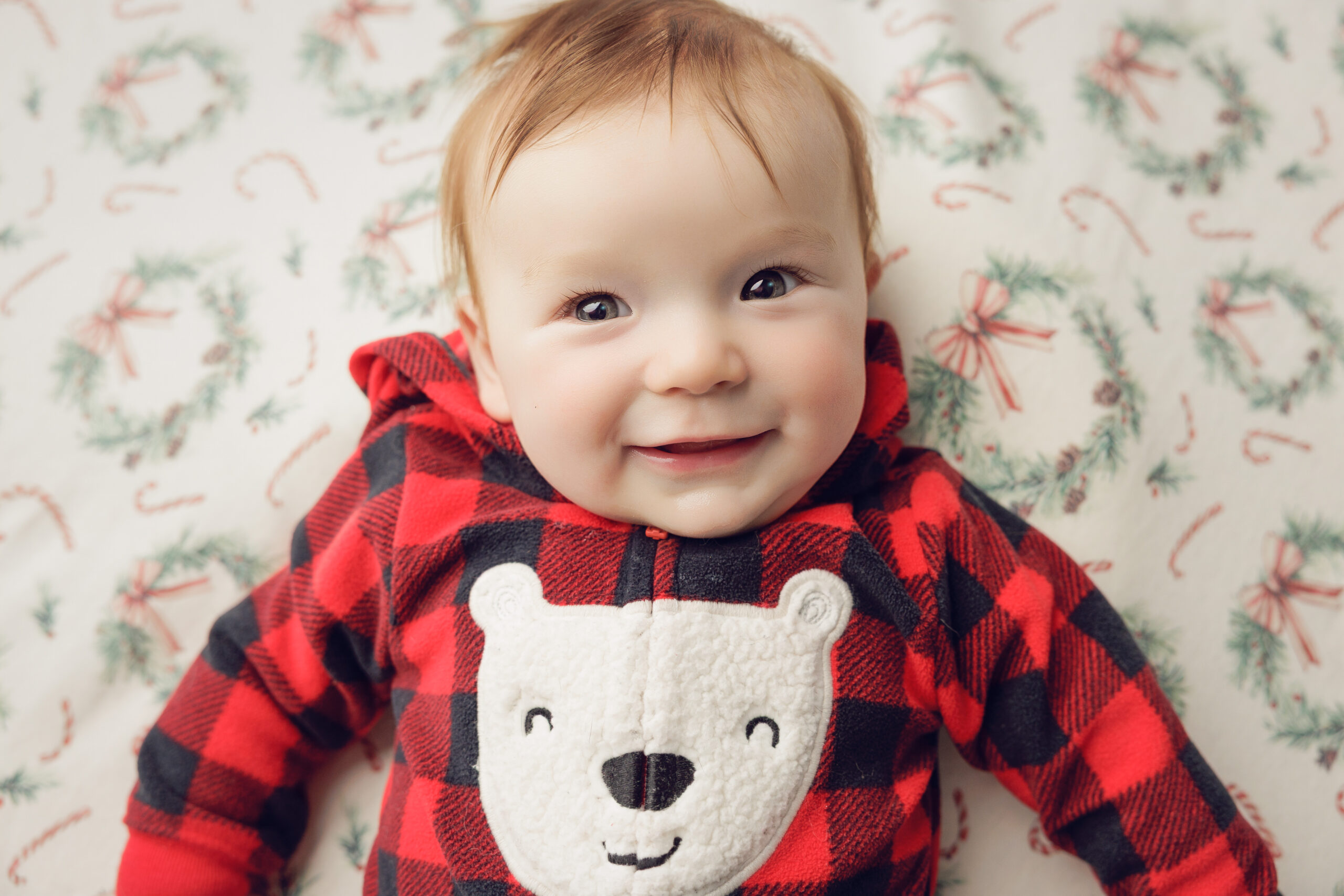 A happy infant lays on a christmas sheet covered bed in red plaid bear pajamas after some mommy and me classes in bloomington il