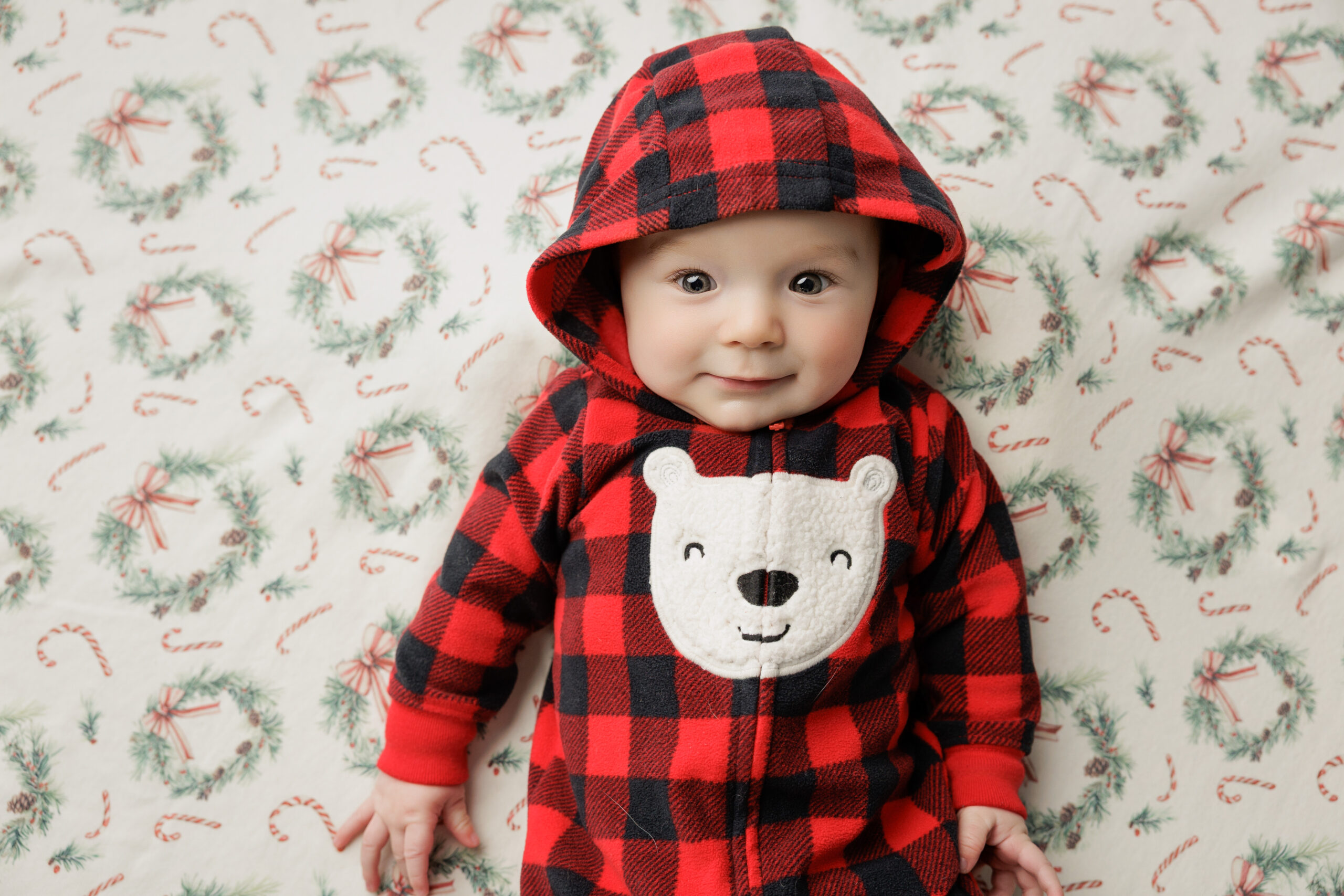 A smiling infant lays on a bed in a red plaid sleep onesie with a hood before enjoying mommy and me classes in bloomington il
