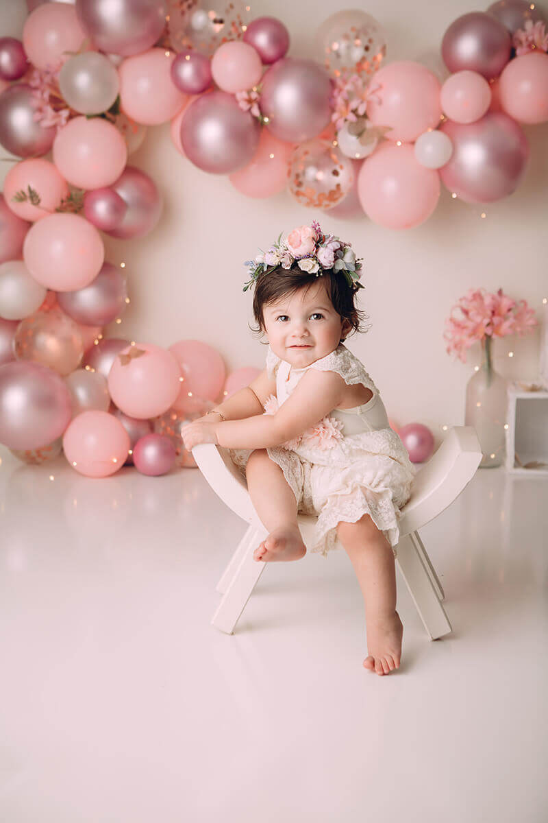 A toddler girl in a pink lace onesie romper plays in a curved stool in a studio