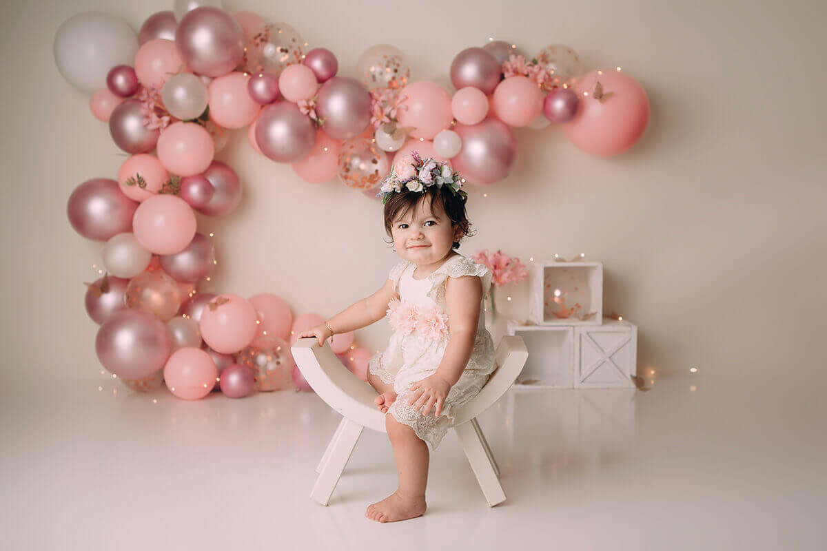 A toddler girl sits on a curved white stool in a studio in a white and pink lace dress after attending montessori children's centre
