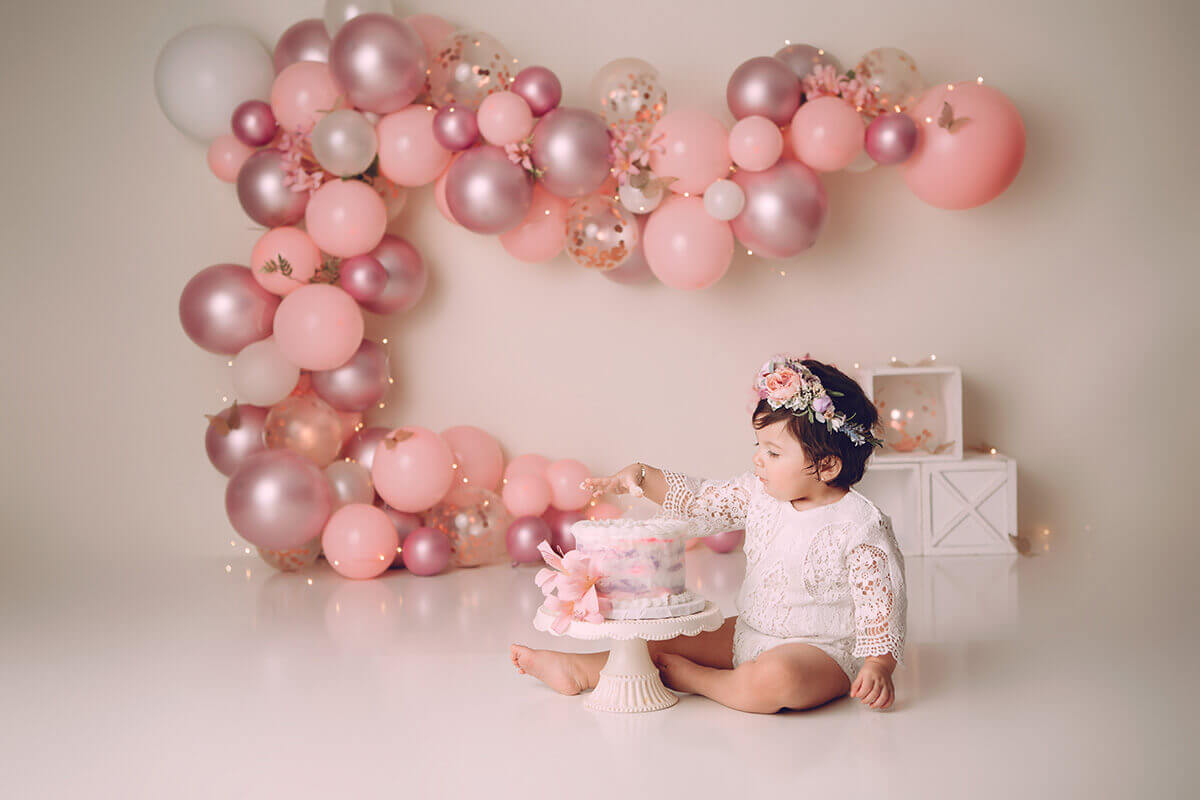 A baby girl in a pink lace onesie pulls her fingers out of a cake in a studio for a cake smash before attending montessori children's centre
