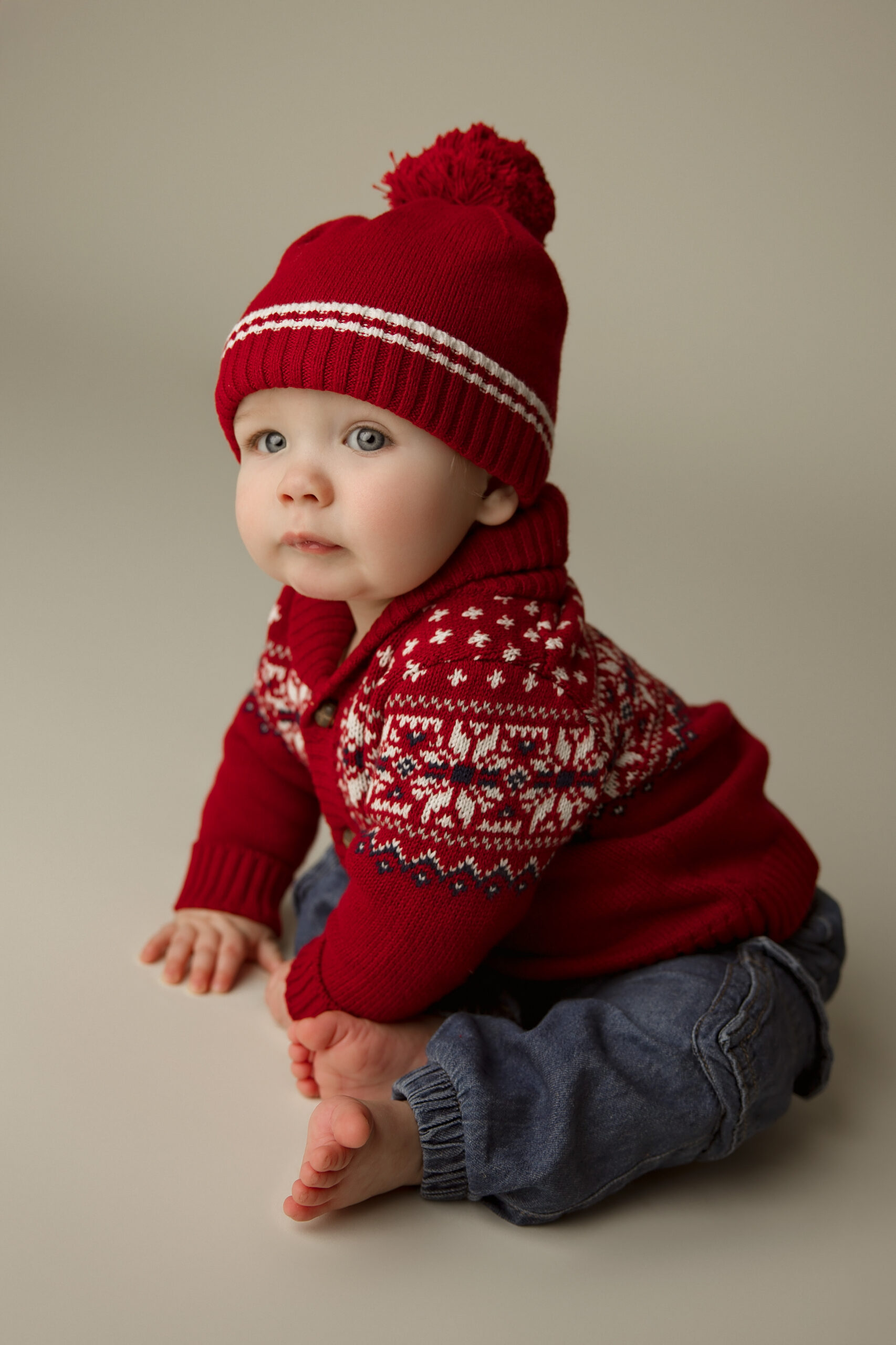A baby sits in jeans and red sweater with matching beanie in a studio