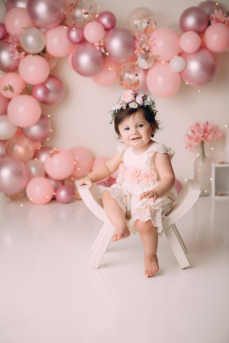 A toddler girl in a pink flower romper sits on a tiny curved stool in a studio in front of pink balloon wall after visiting playgrounds in Bloomington, IL