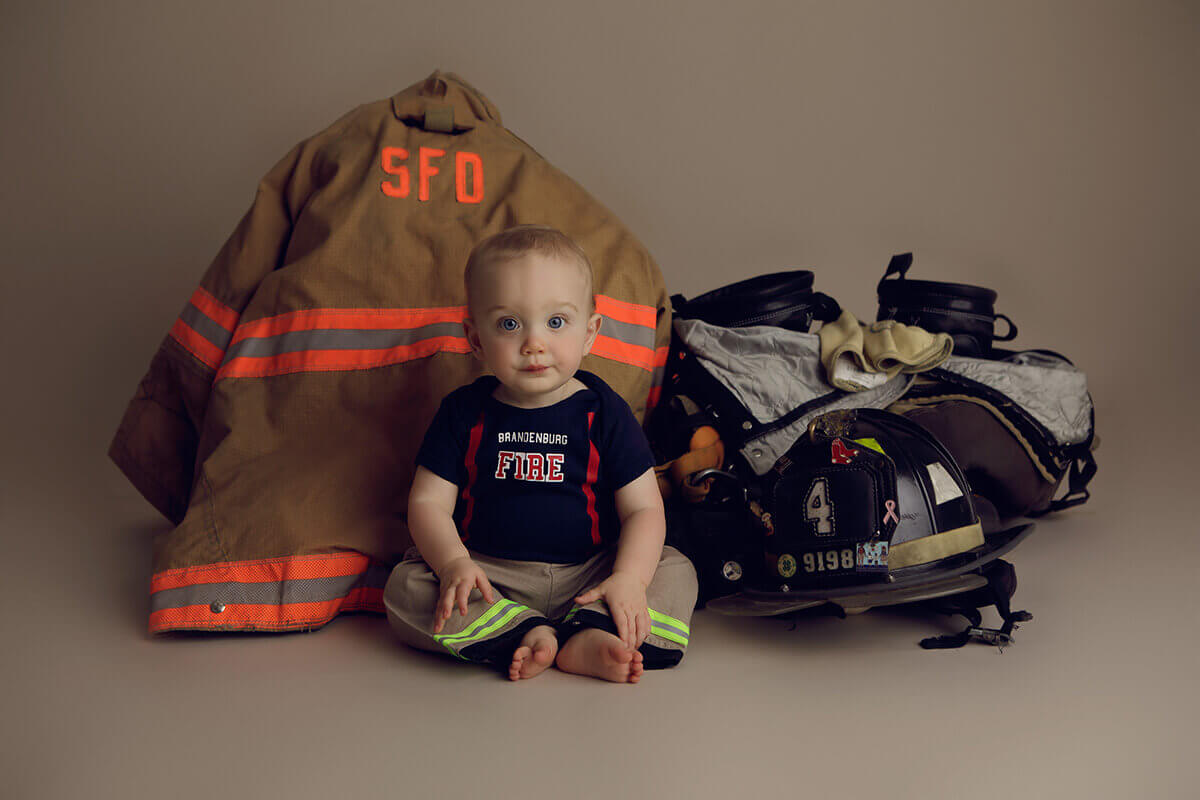 A baby sits in a fire fighter shirt while surrounded by equipment of a fire fighter