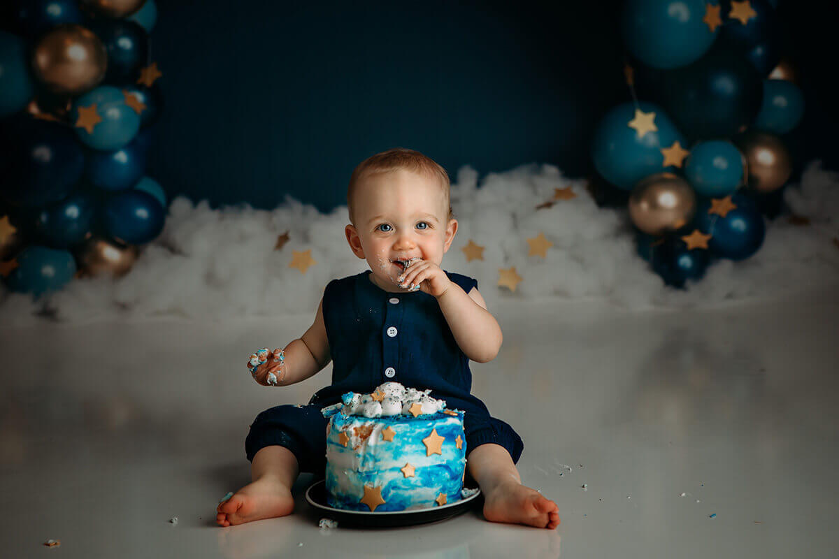 A happy baby sits eating a small blue cake in a studio for a cake smash first birthday after finding preschools in bloomington il