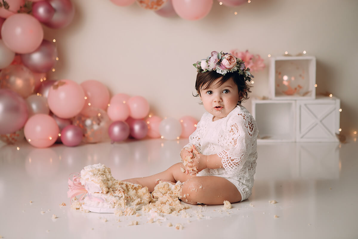 A baby girl sits in her mess after smashing her birthday cake in a white lace onesie and pink rose headband