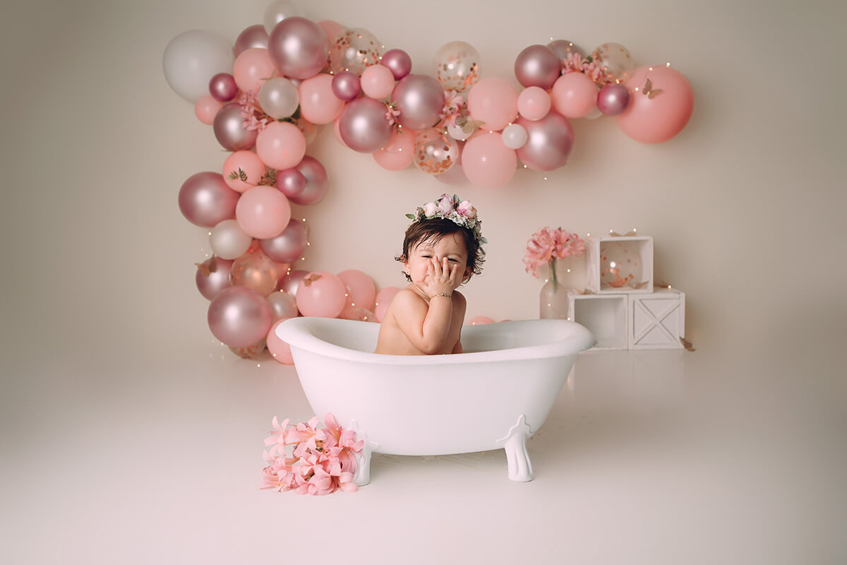 A happy baby girl covers her face with her hand while sitting in a bathtub in a studio with a pink balloon arch