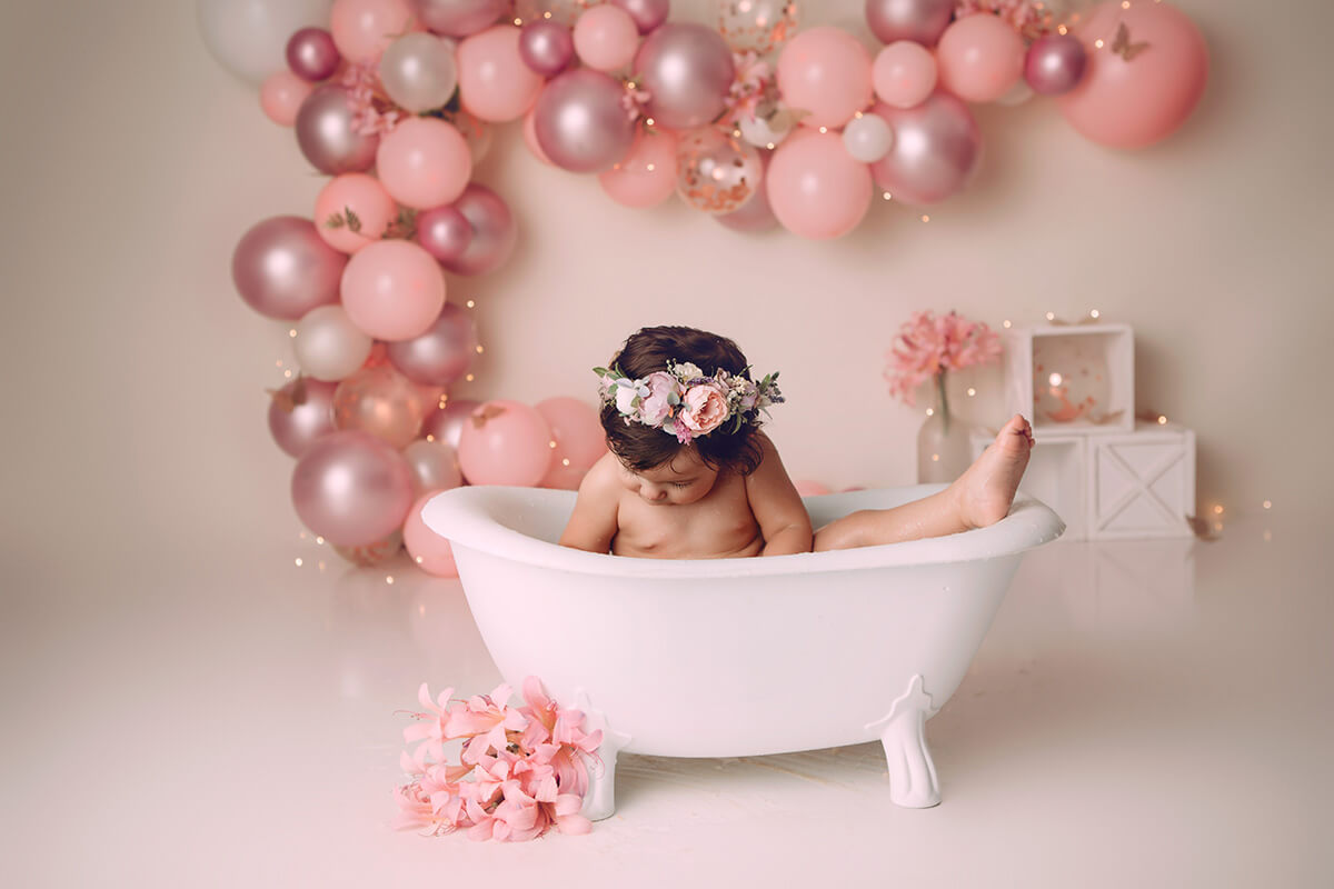 A baby girl sits in a bathtub in a pink studio with a leg sticking out in a rose headband after some swim lessons in bloomington il