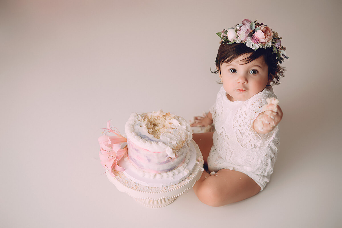 A baby girl in a lace onesie and rose headband offers cake from her birthday in a studio before attending swim lessons in bloomington il