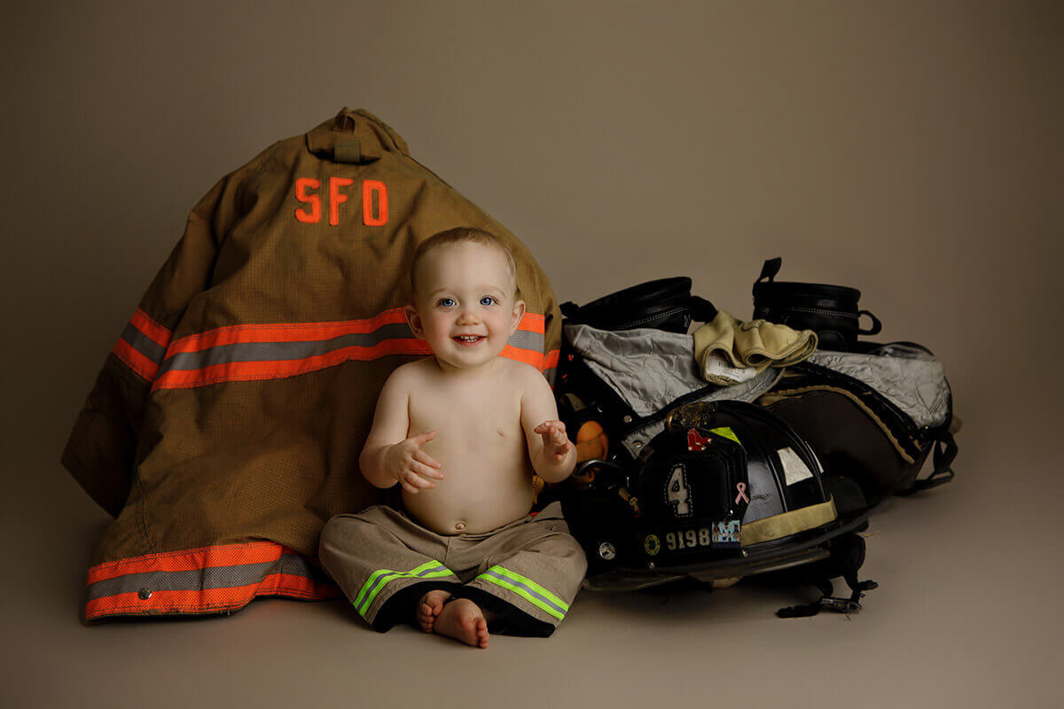 A happy baby boy sits in mini fireman pants surrounded by adult sized fire gear