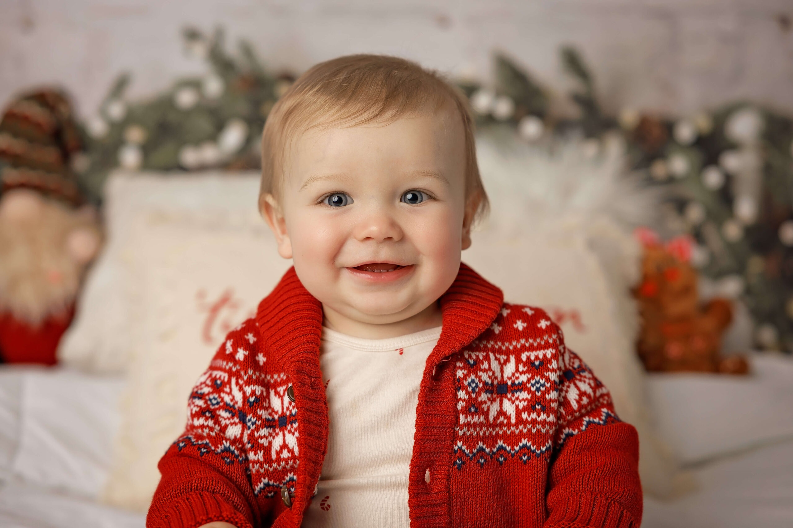 A smiling toddler boy in a red winder knit sweater in a christmas themed studio during some fun things to do with kids in bloomington, IL