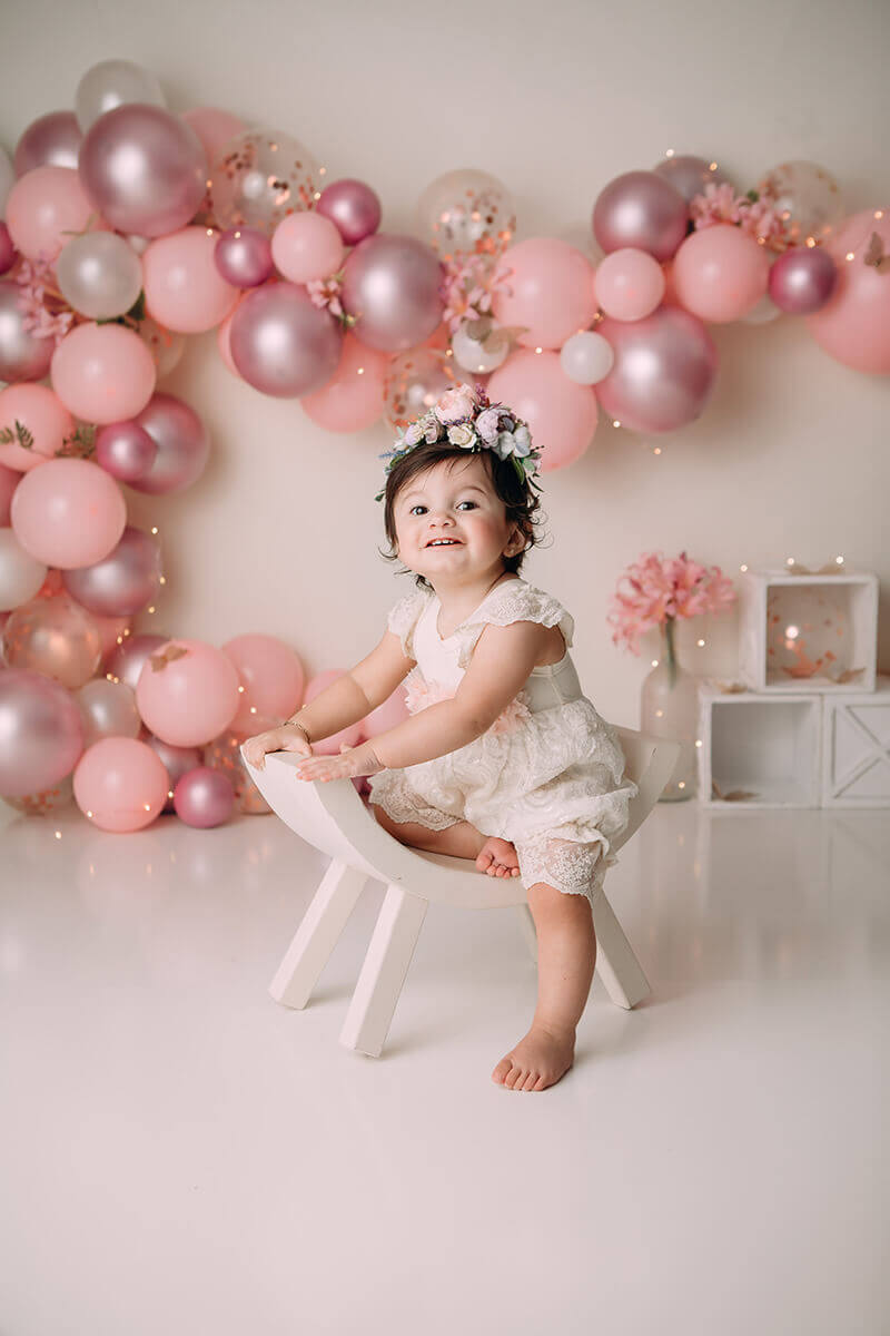 A smiling toddler girl sits in a curved stool in front of a balloon wall in a studio in a pink dress and flower headband