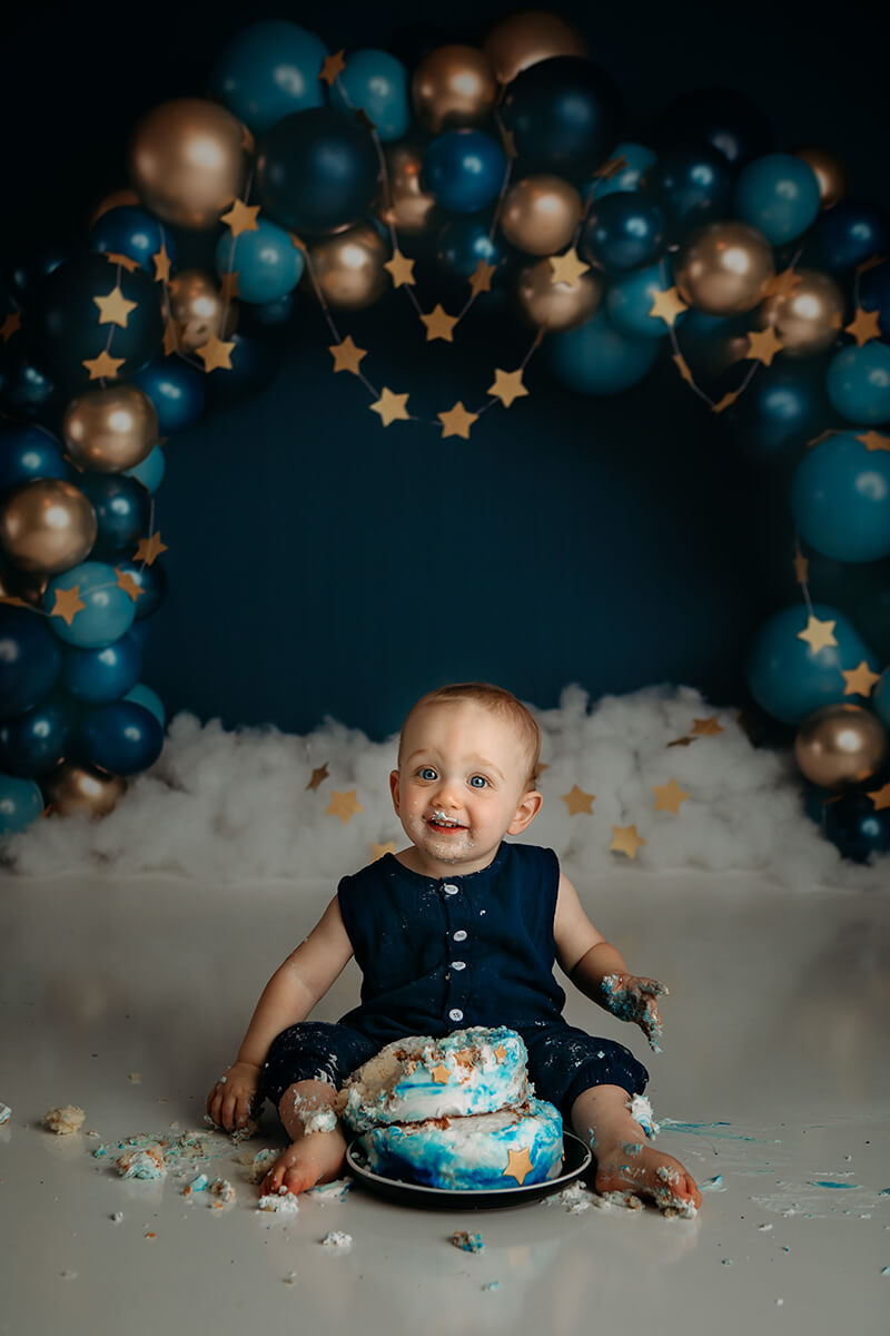 A smiling baby boy sitting in a studio covered in his birthday cake in front of a balloon arch
