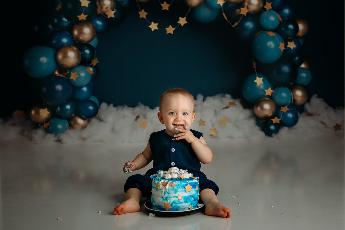 A smiling baby smashes a decorated cake in a blue onesie in a studio after visiting toy stores in bloomington il