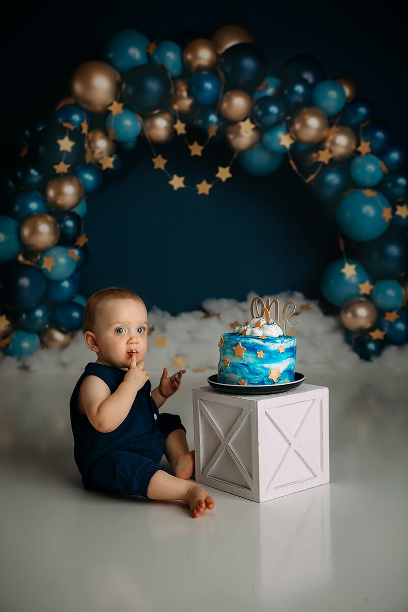 A make eats icing off his finger from a birthday cake on a wooden box in a studio in a blue onesie with balloon arch