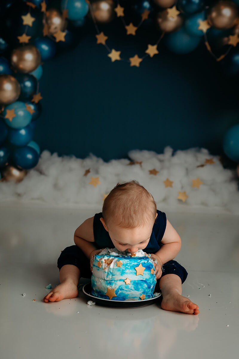 A happy baby sticks his face in a blue cake while sitting in a studio in a matching onesie after visiting toy stores in bloomington il