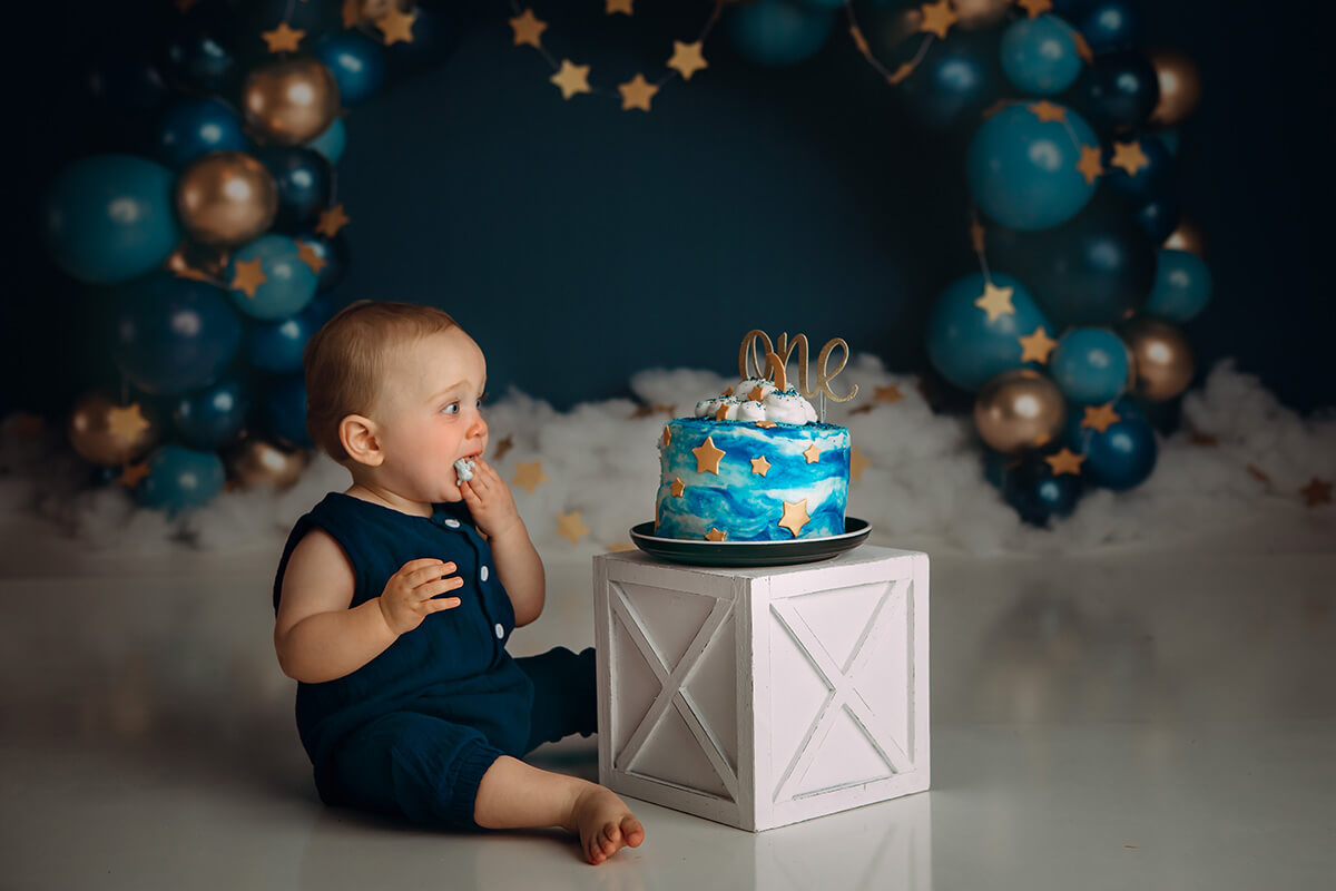 A baby in a blue onesie sits with a sky painted cake for first birthday in a studio after visiting toy stores in bloomington il